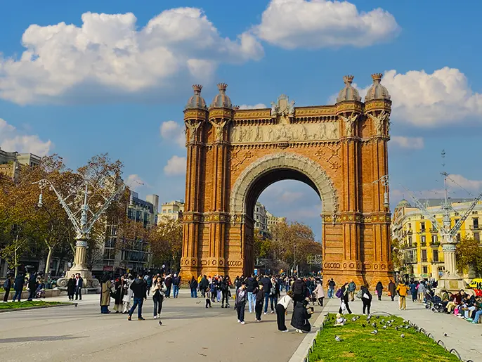 Arc de Triomf (Triumphal Arch) in Barcelona — a large red-brick neoclassical archway with detailed sculptures and coats of arms, serving as the grand entrance to the 1888 Universal Exhibition, surrounded by a busy plaza filled with crowds of people walking and sitting, under a bright blue sky with scattered clouds.