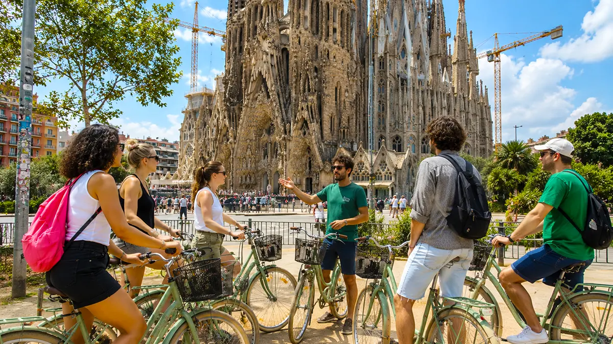 A group of cyclists with their guide stopped in front of the iconic Sagrada Familia during the Barcelona Bike Tour. The guide gestures while explaining the landmark to the smiling participants on their light green bicycles on a sunny day.
