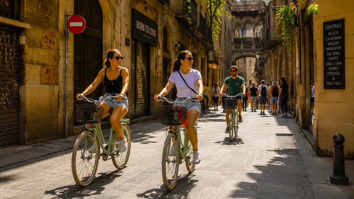 Three happy cyclists riding through the narrow historic streets of Barcelona’s Gothic Quarter on the Barcelona Bike Tour. The women in front smile as they ride past ancient buildings and shops under bright sunlight.