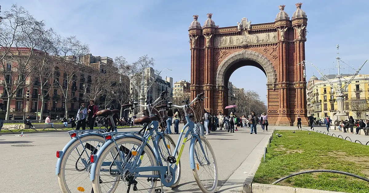 Two light-blue city bikes parked on the promenade leading to the Arc de Triomf in Barcelona, with people strolling and historic buildings lining the sides during Barcelona Bike Tour.