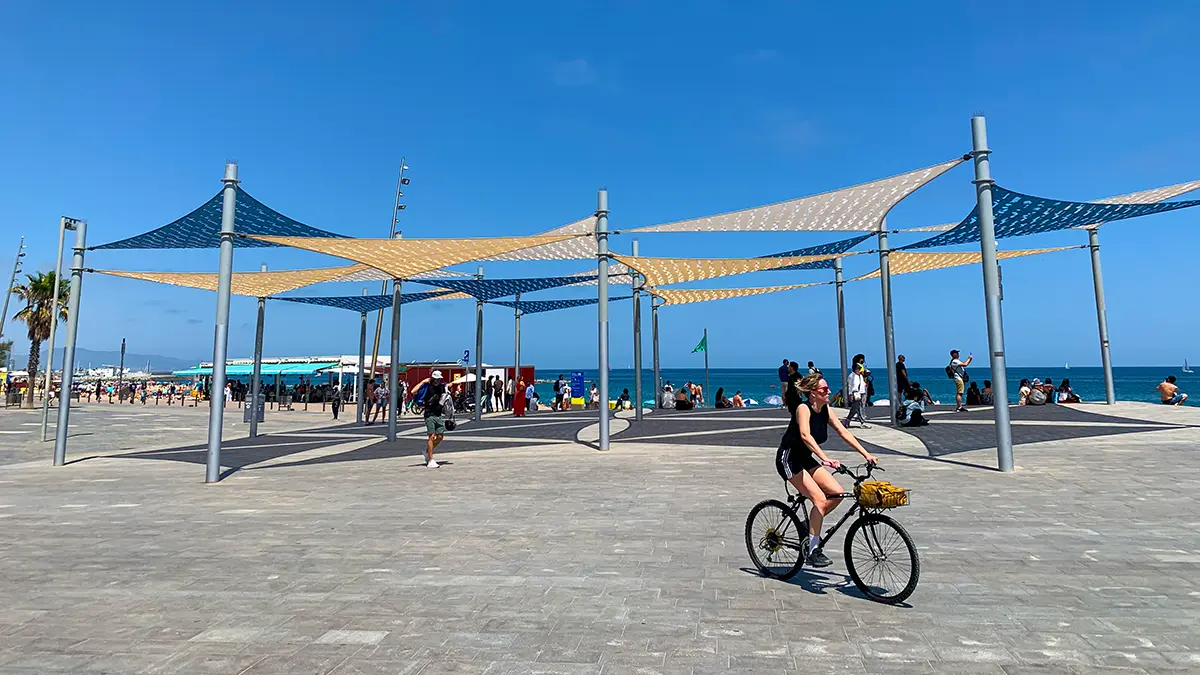 Woman cycling along Barcelona beach promenade under colorful shade sails on the Barcelona Gothic Quarter & Coast Bike Tour.
