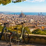Scenic overlook of Barcelona in September with two electric bikes parked by a stone wall, Sagrada Família and the Mediterranean visible across the sunlit city.