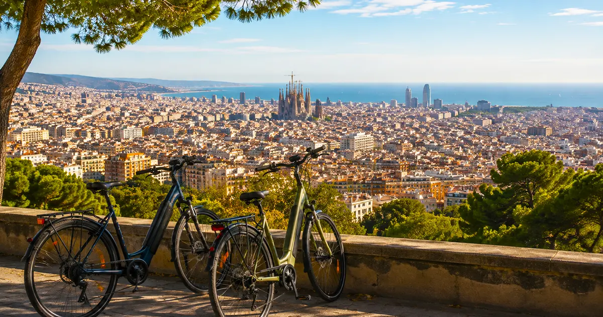 Scenic overlook of Barcelona in September with two electric bikes parked by a stone wall, Sagrada Família and the Mediterranean visible across the sunlit city.