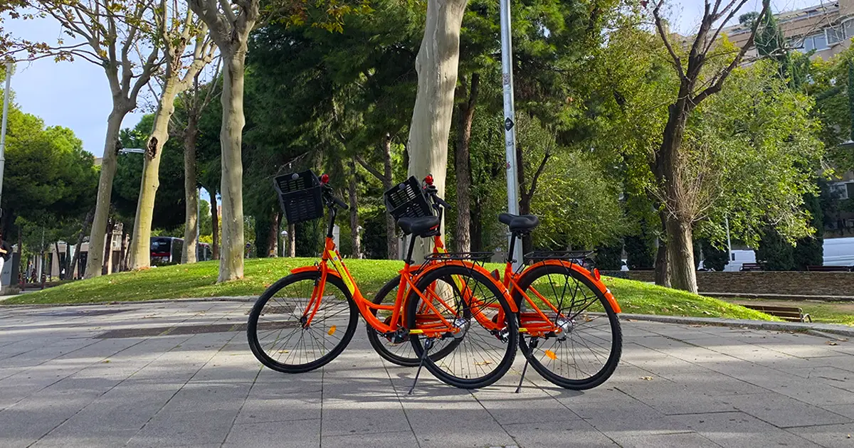 Two bright orange rental bikes parked side by side on a tree-lined sidewalk in a sunny park in Barcelona. The bicycles feature black baskets, comfortable seats, and sturdy frames, ready for exploring the city. Bike rental Barcelona.