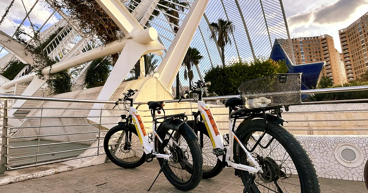 Two white electric fat-tire rental bikes with front baskets and rear racks parked on a modern pedestrian bridge in Valencia, Spain, with the iconic white cable-stayed bridge of the City of Arts and Sciences (likely the Montolivet Bridge) in the background, palm trees, and contemporary buildings under a partly cloudy sky. Bike rental Valencia.