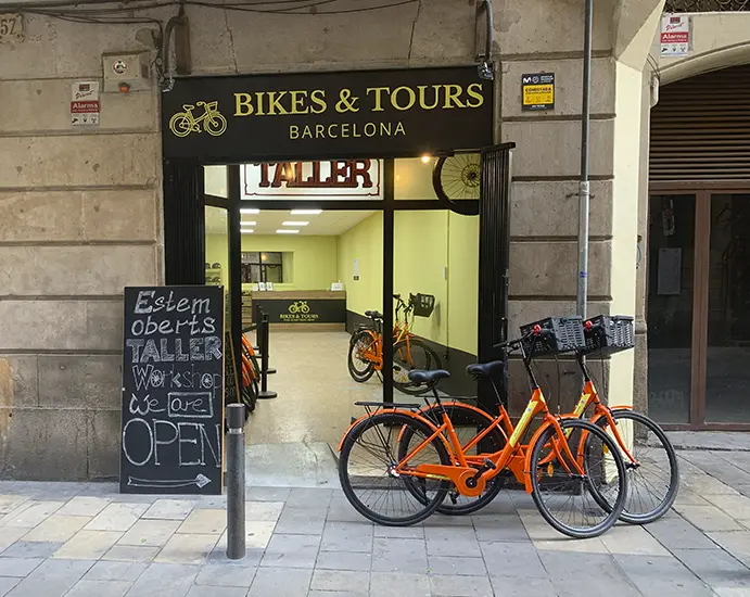 A storefront in Barcelona labeled "Bikes & Tours Barcelona" with a sign above the entrance reading "Taller." Two orange bicycles are parked in front of the open door, and a chalkboard sign on the sidewalk says "Estem oberts Taller Workshop We are OPEN" with an arrow pointing to the entrance. The interior of the shop is visible, showing more bicycles and a counter.