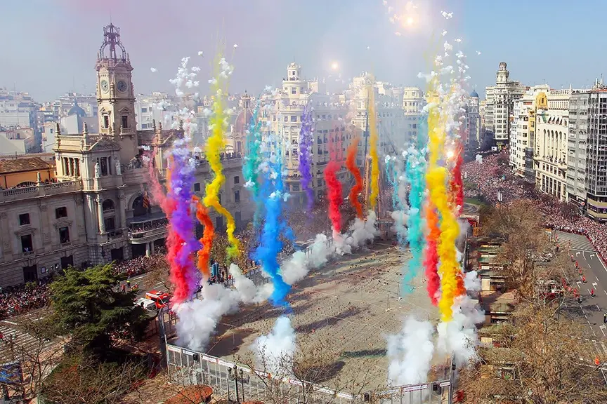 Vibrant aerial view of Fallas Valencia 2026 in Plaza del Ayuntamiento, showing massive colorful smoke plumes in yellow, blue, purple, red, and turquoise rising dramatically from the ground amid a huge cheering crowd, with historic buildings and the city skyline in the background during a mascletà explosion.