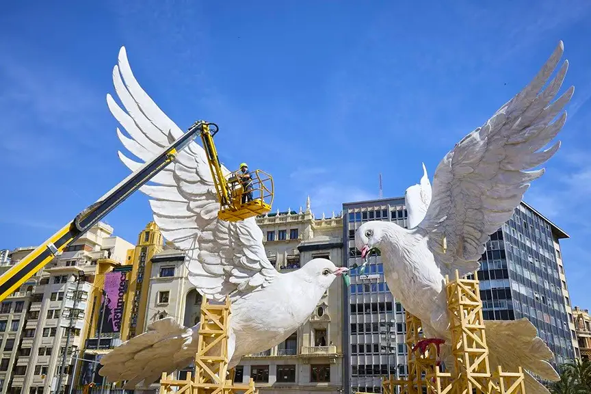 Spectacular daytime setup from Fallas Valencia 2026, showing two enormous white dove ninots with golden beaks on tall scaffolding, workers in a cherry picker connecting them symbolically, set against clear blue skies and modern/valencian architecture in the city center.