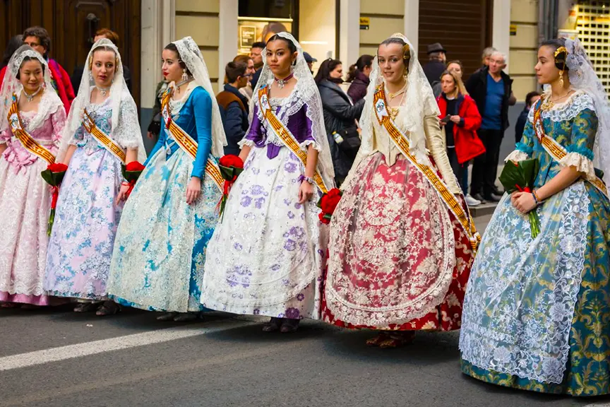Group of elegantly dressed falleras in traditional elaborate gowns during Fallas Valencia 2026, parading in historic Valencia streets; women wearing lace mantillas, colorful silk dresses with floral patterns, sashes, and holding red roses, posing gracefully amid onlookers.
