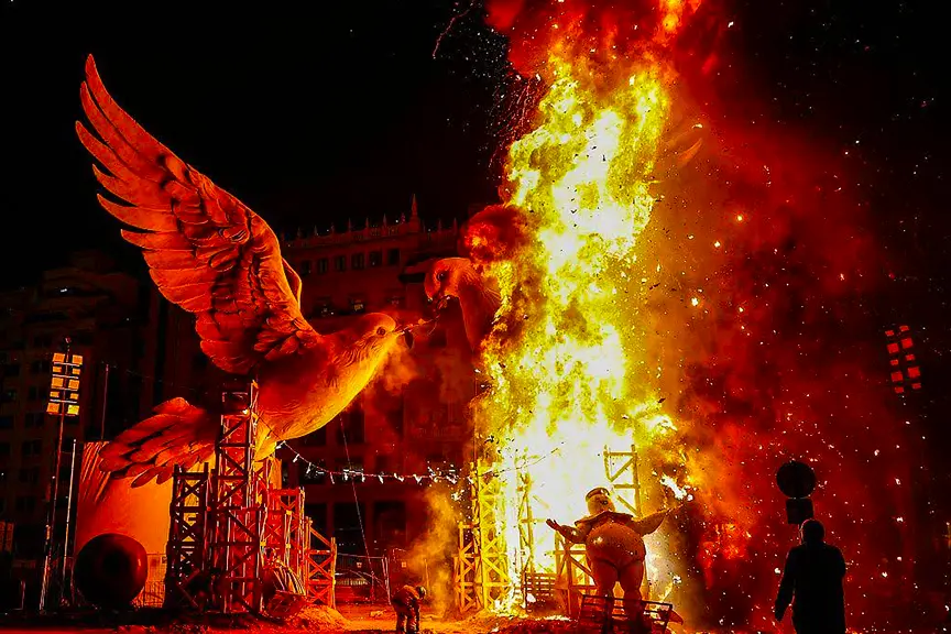 Dramatic nighttime burning at Fallas Valencia 2026, with a large white dove-shaped falla sculpture and a comical oversized piggy-bank figure engulfed in towering bright orange flames, sparks flying against the dark sky and surrounding scaffold structures.