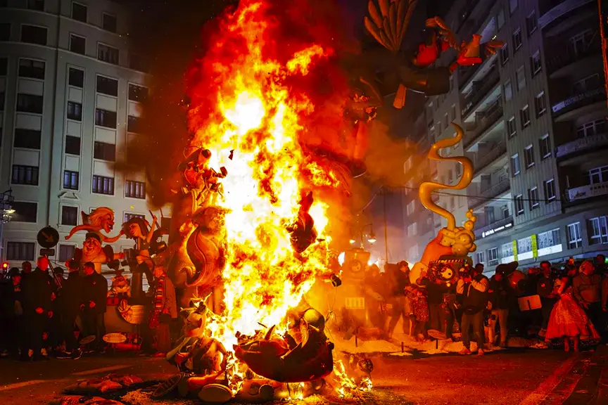 Intense nighttime scene from Fallas Valencia 2026, featuring a towering satirical falla monument engulfed in roaring orange and red flames, with grotesque and fantastical ninots including dragons and mythical creatures burning brightly, surrounded by onlookers and musicians in the street.