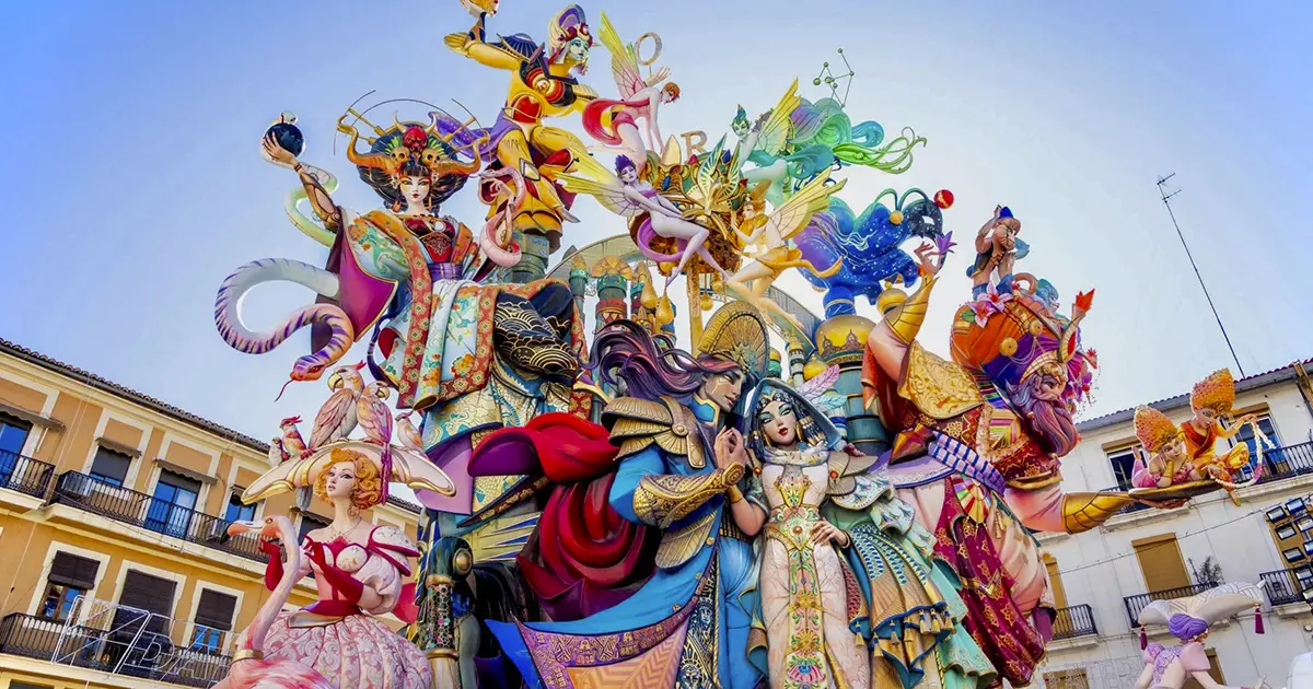 Elaborate towering falla sculpture from Fallas Valencia 2026, showcasing a celestial-themed composition with goddesses, fairies, warriors, colorful serpents, astrological symbols, and ornate costumes in vivid blues, pinks, golds, and reds, displayed in a sunny Valencia square.