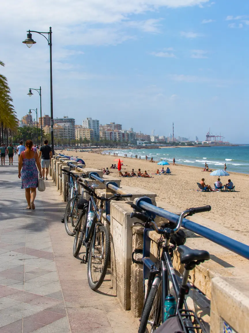 Sunny beach promenade in Valencia lined with bicycles, people walking and relaxing on the sand by the sea, Gay Games Valencia 2026.