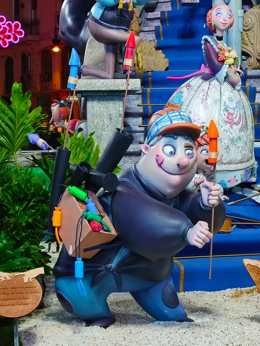 A statue of a cartoon man holding a rocket and a box of fireworks near the plant and a statue of a cartoon woman with flowers behind him.