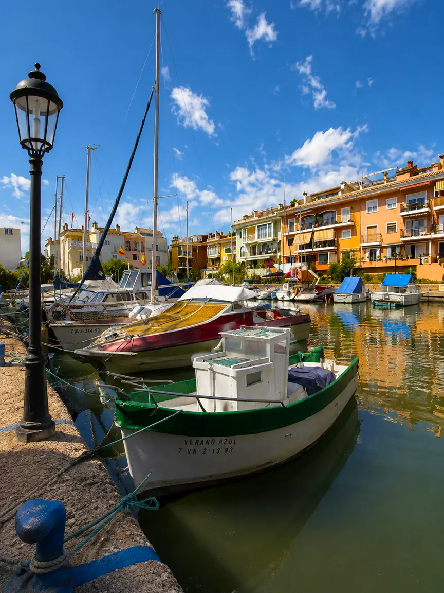 Sunny Port Saplaya harbor view with traditional boats and sailboats docked in Valencia during Macromascletà Universal Valencia 2026, with colorful waterfront buildings in the background.