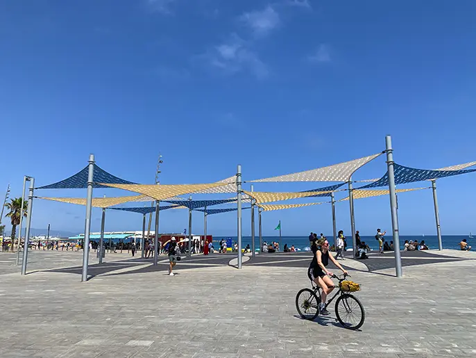 Woman cycling on the sunny Barcelona seafront promenade under colorful shade sails.