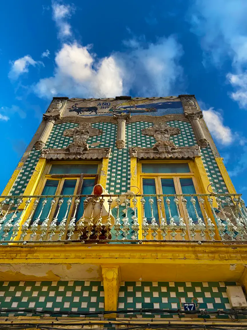 Colorful historic building facade in Port Saplaya featuring green-and-white checkered tiles, yellow window frames, ornate stone decorations, a wrought-iron balcony, and a mosaic panel reading “ANO 1919” against a bright blue sky with clouds.