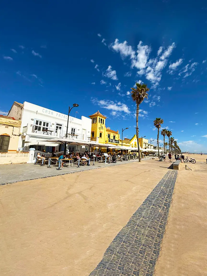 Sunny beachfront promenade in Port Saplaya lined with white and yellow buildings including Casa Patacona, outdoor café seating under umbrellas, tall palm trees, people walking, and a sandy path under a clear blue sky.