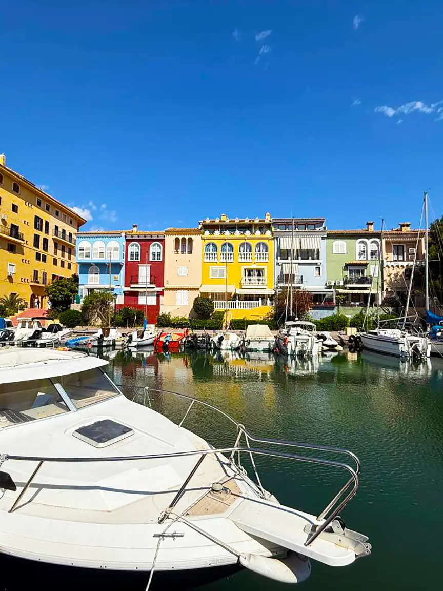 Vibrant Venetian-style colorful houses lining the marina canal in Port Saplaya, with boats docked in calm green water reflecting the buildings under a bright blue sky.