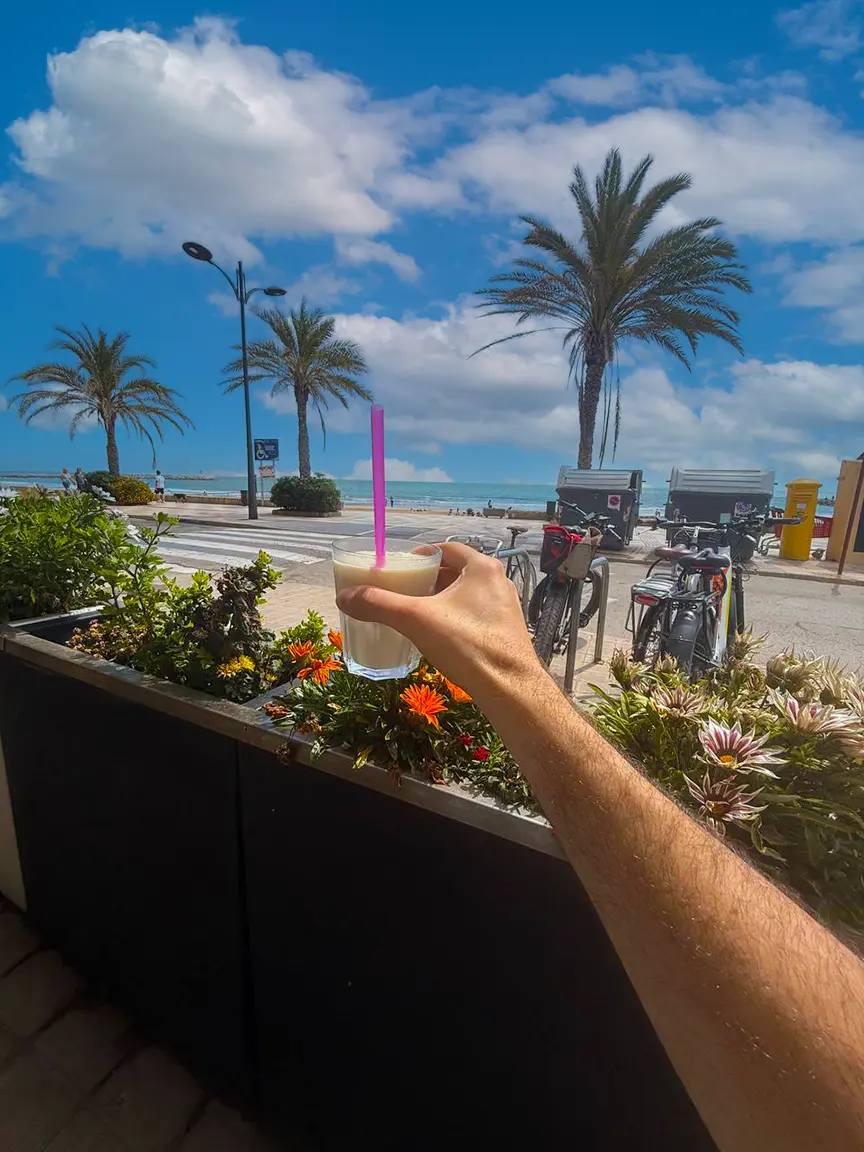 Hand holding a glass of creamy drink with a pink straw in the foreground, overlooking the sunny beach and palm-lined promenade of Port Saplaya with bicycles, flowers, and the sea in the background.