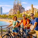 Happy group of three friends smiling at the camera while riding bikes together on the wide beachfront bike route in Barcelona, with Sagrada Família basilica and golden beach in the background.