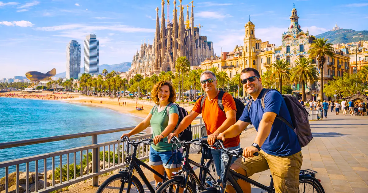 Happy group of three friends smiling at the camera while riding bikes together on the wide beachfront bike route in Barcelona, with Sagrada Família basilica and golden beach in the background.