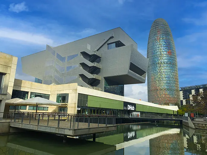 Disseny Hub Barcelona museum beside a reflecting pool, with the cylindrical glass Torre Glòries skyscraper rising behind against a blue sky.