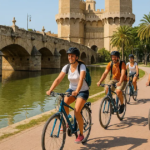 A group of people cycling along a path beside a canal with a historic bridge and tower in the background, sharing Valencia cycling tips.