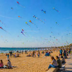 Hundreds of colorful kites flying over crowded beach at Valencia Kite Festival 2026, with palm trees and festival-goers enjoying the event