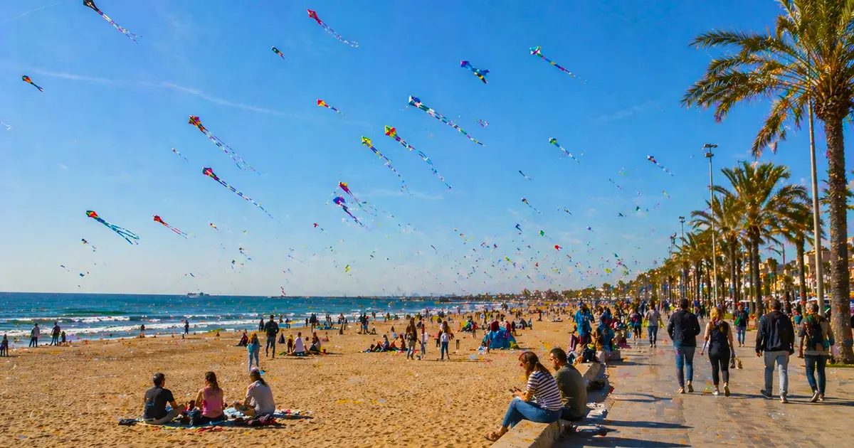 Hundreds of colorful kites flying over crowded beach at Valencia Kite Festival 2026, with palm trees and festival-goers enjoying the event